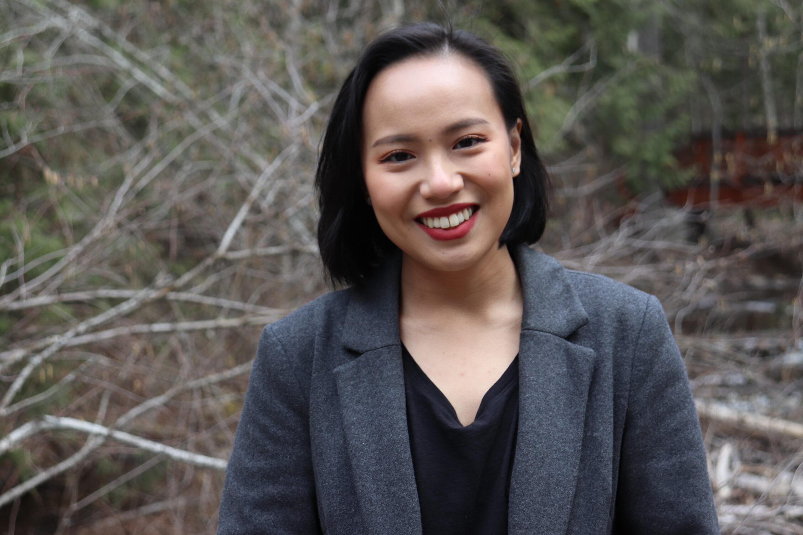 Headshot of Twila Amato, who's wearing a black top, grey blazer, and standing in nature.