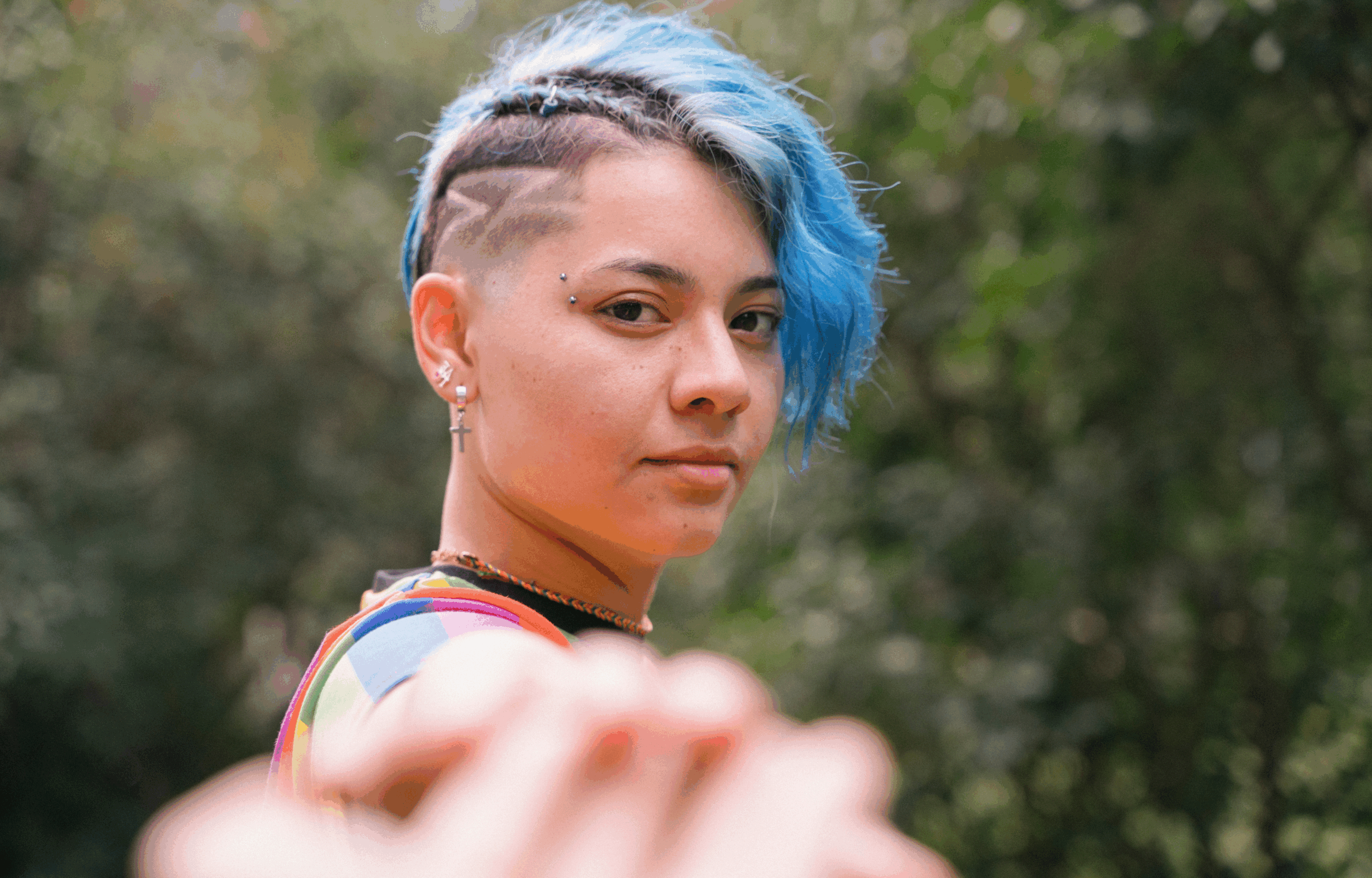 Youth with blue hair and shaved head and rainbow shirt pointing at camera