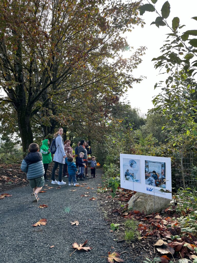 A group of small children and caregivers walk the Sq’ewqéyl I:mex Xalh (Skowkale Walking Path) and there's a sign with cultural stories