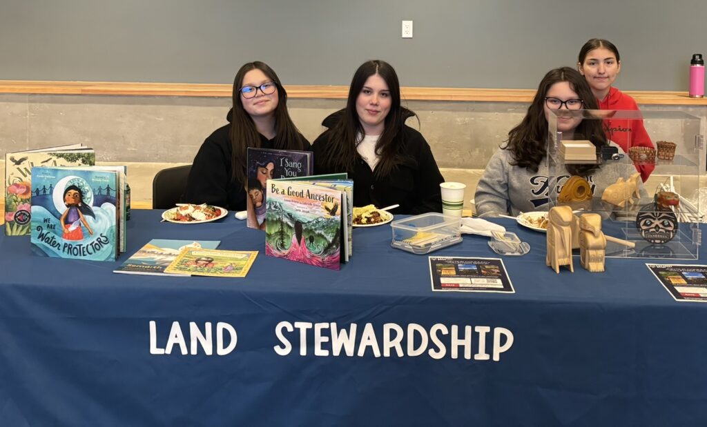 Four young women smiling behind table with books on it and a sign that says Land Stewardship