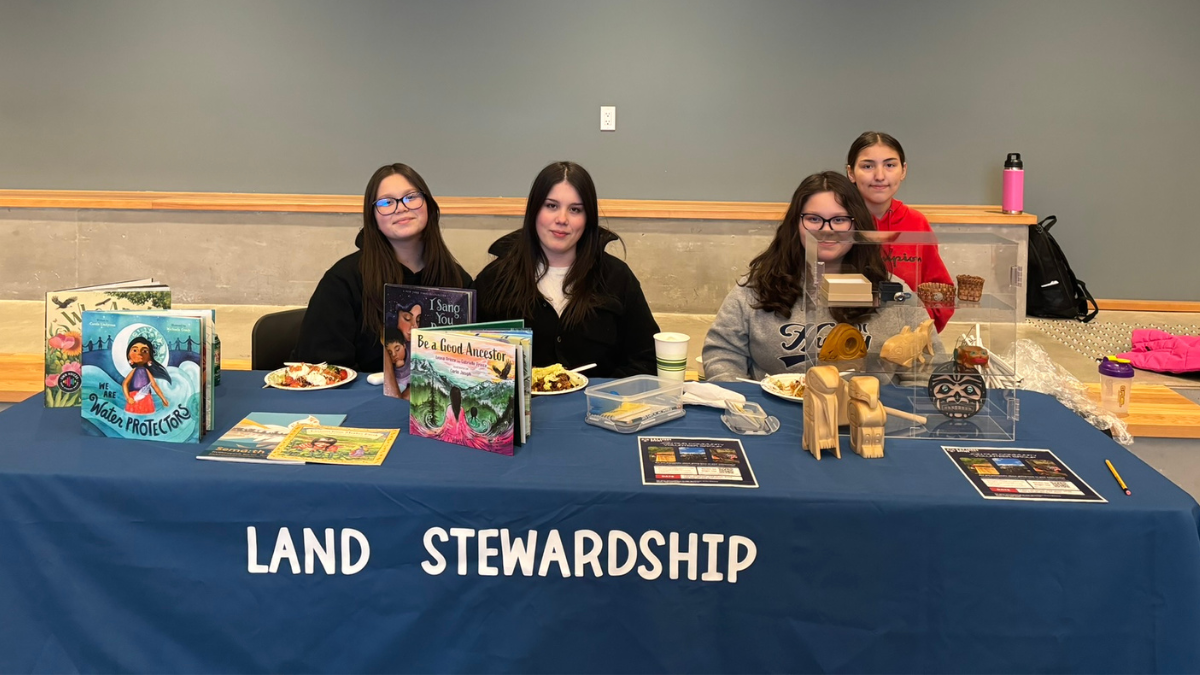 Four young women smiling behind table with books on it and a sign that says Land Stewardship